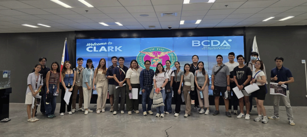 A group of PA 152 MWV students stands inside the BCDA office at Filinvest Innovation Park, holding
printed maps given to them for their field visit to New Clark City. Behind them is a large screen displaying
a welcome message from the Bases Conversion and Development Authority. The students are smiling
and standing closely together, and the modern, well-lit interior of the office reflects a professional and
welcoming environment.